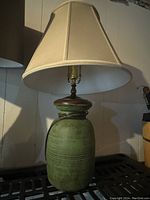 Photo of green ceramic table lamp with beige shade on a dark surface against light-colored wall.