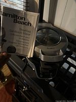 Hamilton Beach Breakfast Sandwich Maker appliance shown with user manual in hand. Device is silver with black handle, designed for making breakfast sandwiches. Visible 'HOT SURFACE' warning on handle.