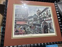 Framed print of a German street scene in Rudesheim with visible signature and handwritten title, detailed architecture and people in outdoor market and cafes.