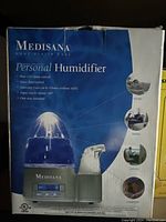 Box showing Medisana Personal Humidifier product features including blue LED lamp control, vapor flow control, automatic shut off after 8-9 hours, vapor noise reduction, and one-year warranty. Highlights use for home, travel, office, comfort.