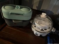 Photo showing two small kitchen appliances for egg preparation: a collapsible electric steamer with a green lid and a white electric egg cooker with a transparent lid.