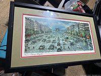 Framed, matted Parisian street scene depicting the bustling Avenue des Champs-Élysées with the Arc de Triomphe under a blue sky with clouds. Multiple cars and pedestrians in the foreground.