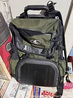 Front view of the Pelican backpack showing olive green fabric, black hardcase front pocket, and zippered compartments.