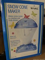 Photo of the box showing the Rival Snow Cone Maker product with features listed: makes snow cones, icy slushes, fruity drinks, soft fluffy snow, uses standard ice cubes from freezer with no special freezing mold required.