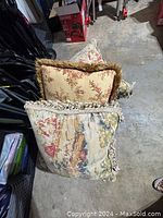 Three decorative throw pillows stacked, showing floral and tapestry designs with fringed and ruffled trims.