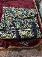 Closed view of the Vera Bradley garment bag folded, showing primary paisley pattern in blue and green and black carrying straps with a flap closure.
