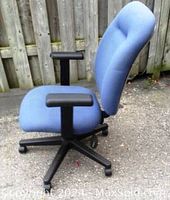 Side view showing dark blue padded seat and backrest with black plastic arms and legs, set outdoors on pavement with wooden fence background.