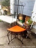 Front left angled view of metal corner baker's rack showing orange wooden bottom shelf and metal middle and top shelves with decorative metal frame