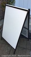 Folding sandwich board standing outdoors with white vinyl panel inserted on one side, black metal frame visible.