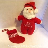 Front view of the Santa stuffed toy with red suit and "My First Christmas" embroidery, sitting next to small red stocking with white snowflake design, both with hanging loops.