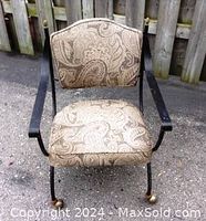 Front view of one wrought iron chair with paisley upholstered seat and back, curved armrests, metal frame, and wheels on legs.