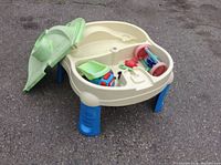 Beige plastic outdoor sand and water table with blue legs, green half lid open, showing play accessories inside.