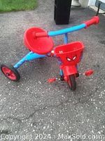 Front and left side view of the red and blue toddler tricycle showing the Spiderman logo on the front basket, red seat, handlebars, and wheels.