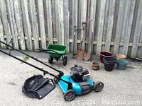 View of the lawn mower, wheelbarrow, garden tools including two shovels, rake, and plant pots arranged against a fence.