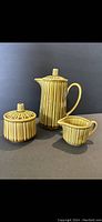 Front angle view of the mustard colored 3 piece ceramic tea set showing teapot, creamer, and sugar bowl with lids, displaying ribbed vertical lines and lids' knob handles.