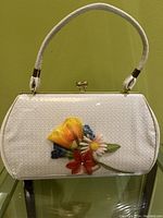 Front view of vintage white plastic purse with colorful embedded flowers behind plastic and white patent handle, showing clasp closure.