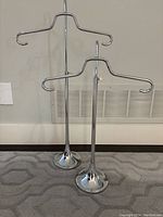 Two chrome expandable display hangers, one in front and one slightly behind, both standing on carpeted floor.