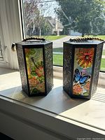Two patio lanterns with hexagonal shape, black metal frames and colorful stained glass panels showing flowers and butterflies, braided rope handles on top, placed on window sill