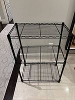 Front view of black metal 3-tier shelving unit showing wire grid shelves and vertical posts.