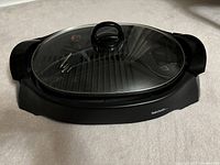 Front view of the Betty Crocker electric grill assembled with glass lid in place, showing the black ridged cooking surface beneath the clear lid and black body with handles.