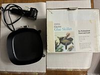 7-inch square black electric glue skillet, showing surface, temperature dial on side, detachable electrical plug, and original box with label "Electric Glue Skillet" for floral and craft use.
