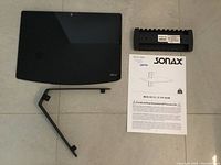 Top view of the black glass shelf, metal bracket, plastic supports, product manual, and Sonax label.