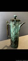 Side view of tall pottery coffee urn showing textured circular pattern glaze in turquoise and dark brown.