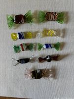Nine assorted multi-coloured Murano glass candy figurines arranged on a white surface, showing variety in color and design.