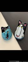 Photo showing two Picasso style cat-shaped ceramic salt and pepper shakers, one turquoise and one white/black, standing on a flat surface.