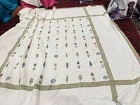 Full view of large cream-colored coverlet with a grid of embroidered flowers and olive green borders.