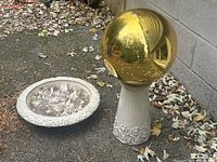 Overall view of bird bath components showing gold sphere on pottery stand next to detached bird bath bowl on the ground with leaves around.