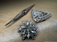 Front view of three brooches on wooden surface showing Victorian marcasite elongated piece, triangular rhinestone piece, and round starburst rhinestone piece.