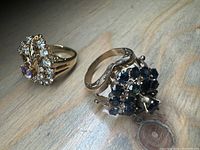 Photo showing two gold plated rings on a wooden surface, one with clear and purple synthetic stones and one with dark blue synthetic stones.