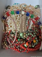 Top view of the box filled with tangled costume jewelry including pearl-like beads, colorful plastic beads in various shapes and sizes, and some metal chain accents