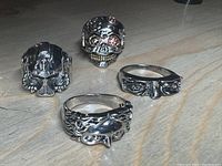 Four silver-tone rings on a wood surface showing various artistic designs including faces, eyes, a skull, and a snake.