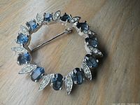 Close-up photo showing the circular brooch with blue oval rhinestones alternating with clear marquise rhinestones on a silver-tone backing.