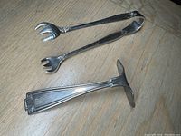 Two sterling silver serving pieces on wooden surface: a food pusher and a pair of olive/pickle tongs with floral decorations at handles.