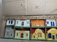 View of eight chalkware Christmas village buildings arranged in original box, showing size scale with ruler. Colors include beige, orange, red, yellow, and white with painted details. Original cellophane window covering partially intact.