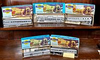 Five unopened Athearn HO scale Santa Fe Silver passenger car kits displayed on a wooden shelf. Boxes show vintage artwork with a steam train and mountain scenery, cars inside are silver passenger coaches and baggage car models.