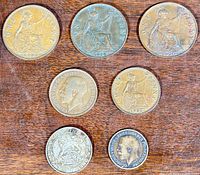 Seven old British coins laid flat on a wooden surface with visible dates and Britannia design on pennies and half pennies, and crowned lion and portrait on shillings.