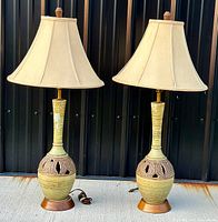 Pair of vintage mid-century modern ceramic table lamps with matching cream fabric lampshades on a concrete floor with a black metal wall backdrop.