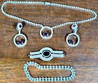 Four-piece vintage rhinestone demi parure set with amethyst-colored center stones displayed on wooden surface: necklace, earrings, bar brooch, and double row bracelet.