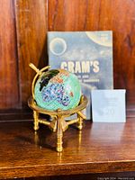 Photo showing the globe with multiple semi-precious stones visible, brass stand, and the blue handbook behind