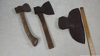 Lot displaying two small axes with wooden handles and one large single bevel axe head with no handle; laid out next to a ruler for size comparison.
