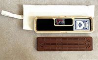 Wooden cribbage board with playing cards and scoring pegs inside a wooden tray, displayed next to a white fabric carrying case.