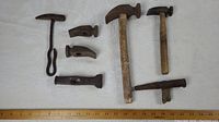 View of seven drop forged hammer heads and hammers arranged on white surface with ruler for scale. Mixture of hammer heads and two complete wooden handled hammers visible.
