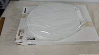 Full view of the white toilet seat in plastic wrap on top of the Kohler branded box.
