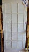 Full front view of the white door with 15 glass panes in grid layout, partially wrapped in clear plastic film, showing size and brand tag.