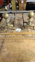 Full pair of vintage brass and iron fireplace andirons placed side by side on a wooden surface.