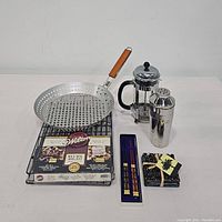Lot laid out showing all items: perforated BBQ skillet with wooden handle, glass French press, metal cocktail shaker, new Wilton cooling racks, coaster set, and chopsticks.